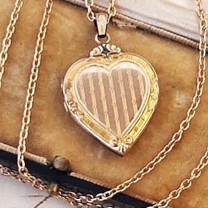 May include: A gold-toned heart-shaped locket pendant suspended from a matching link chain. The locket features a central panel with vertical engraved stripes and a decorative border with ornate scrollwork details along the outer edges.