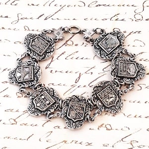 May include: A silver-toned link bracelet featuring six ornate shield-shaped charms. Each charm displays a unique crest and the name of a French city: Caen, Lisieux, Le Havre, Rouen, Bagnoles, Cherbourg, and Mont Saint-Michel, connected by metal links.