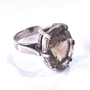 May include: A silver ring featuring a large, oval-cut, translucent brown gemstone held in place by six prongs. The metal band has a smooth, polished finish and connects to the setting with a structured, geometric design on both sides of the stone.
