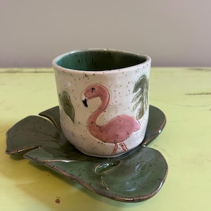 May include: A ceramic cup and saucer set. The cup is white with a pink flamingo design and green leaf accents. The saucer is leaf-shaped and green. The cup and saucer are speckled with brown dots.
