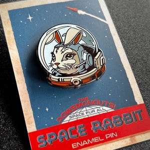 May include: A red, white, and blue enamel pin featuring a cartoon rabbit wearing an astronaut helmet. The pin says "The Dawnsmonouts! Space Fur All" and "Space Rabbit Enamel Pin".