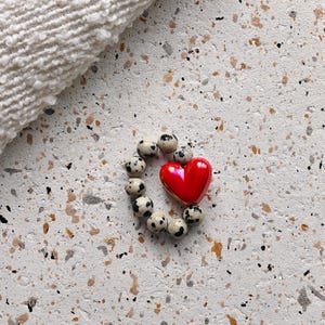 Könnte beinhalten: Ein Kreis aus kleinen, gesprenkelten Perlen umgibt eine glänzende, rote herzförmige Perle. Die Perlen sind auf einer gesprenkelten, hellfarbigen Oberfläche angeordnet, mit einem weißen, strukturierten Stoff in der oberen linken Ecke.