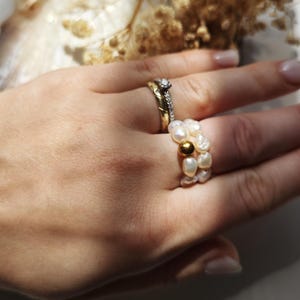 Könnte beinhalten: Eine Hand mit zwei goldenen Ringen und einem Perlenring. Die goldenen Ringe haben einen Diamanten und ein strukturiertes Band. Der Perlenring besteht aus weißen Perlen und einer goldenen Perle.