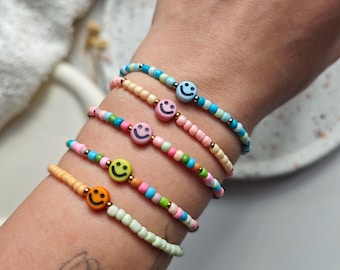 May include: Five beaded bracelets with smiley face charms. The bracelets feature a variety of colours, including pink, blue, green, and yellow. Each bracelet has a different colour scheme and a gold-coloured bead.