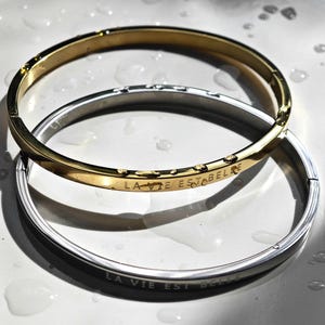 Könnte beinhalten: Zwei aufklappbare Armreifen, einer in Goldoptik und einer in Silberoptik, jeweils mit der Gravur LA VIE EST BELLE versehen. Die Armreifen haben eine glatte, polierte Oberfläche und liegen übereinander, wobei kleine Wassertropfen zu sehen sind.