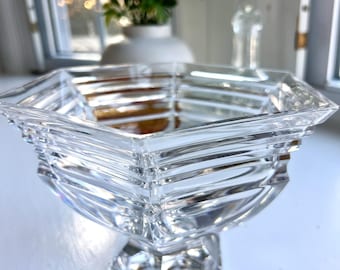 May include: A clear, faceted crystal bowl with a square shape and a pedestal base. The bowl has a geometric design with horizontal and vertical lines, creating a textured appearance. The bowl is sitting on a white surface.