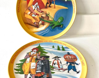May include: Two vintage McDonald's plates with yellow rims. One plate depicts Ronald McDonald on a beach with an octopus, the other shows Ronald and the Hamburglar in a snowy scene with a snowman. Text includes "Ronald McDonald saves this hot summer day..." and "Ronald McDonald and Hamburglar share a big grin..."