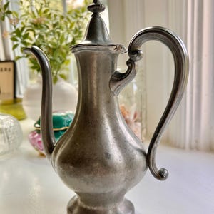 May include: Antique silver-colored metal pitcher with a curved handle and a spout. The pitcher has a decorative lid with a small finial. The base of the pitcher is round and sits on a small pedestal. The pitcher is likely used for serving beverages.