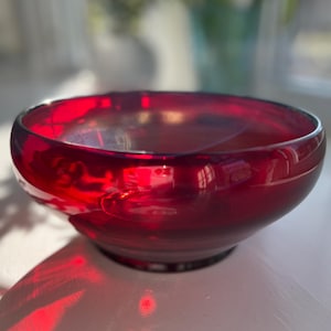 May include: A round, ruby red glass bowl. The bowl has a smooth, glossy finish and is sitting on a white surface. The light reflects off the bowl, creating a bright, vibrant appearance. The bowl is empty and appears to be a decorative item.