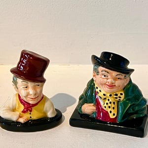 May include: Two ceramic figurines depicting gentlemen. One wears a burgundy top hat and yellow vest, the other a black hat and green coat. Both have black bases and are painted with detailed facial features.