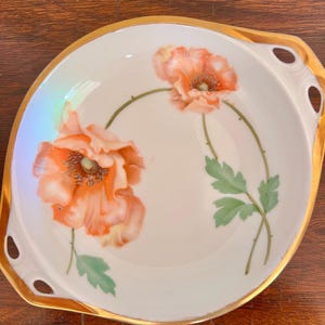 May include: A white porcelain serving dish with a gold rim and handles. The dish features two orange poppy flowers with green leaves and stems. The flowers are painted in detail, with a light orange and yellow gradient.