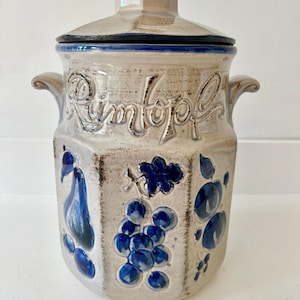 May include: A beige ceramic Rumtopf jar with a lid, featuring blue fruit designs and the word "Rumtopf" embossed on the front. The jar has two handles and a blue rim.