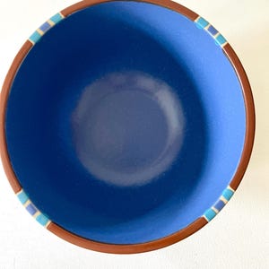 May include: A deep blue ceramic bowl with a brown rim. The rim features a decorative band of small blue, teal, and beige squares. The bowl's interior has a lighter blue hue, creating a contrast with the darker exterior.