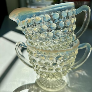 May include: A stack of two opalescent glass pieces featuring a raised hobnail texture. The top piece is a creamer with a pouring spout and a single handle, while the bottom piece is a sugar bowl with two side handles and a pedestal base.