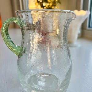 May include: A clear glass pitcher with a green handle. The pitcher has a rounded body and a spout for pouring. The glass has a slightly bubbly texture. The handle is a lighter shade of green than the body of the pitcher.