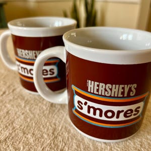 May include: Two brown ceramic mugs with white rims and handles. The mugs feature the Hershey's S'mores logo in white, orange, and blue. The mugs are on a beige surface.