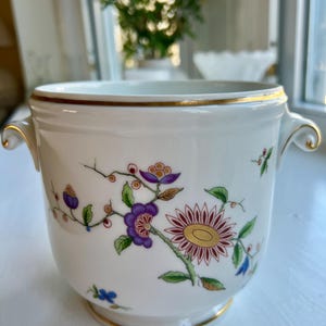 May include: A white porcelain bowl with gold trim and handles. The bowl is decorated with a floral pattern featuring purple, red, and yellow flowers with green leaves. The bowl has a slightly flared base.