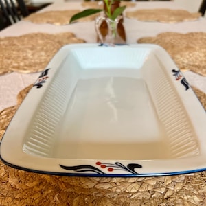 May include: A rectangular white serving dish with a blue trim and decorative floral accents. The dish has a ribbed interior and is set on a woven placemat. The floral design includes red berries and black and blue curved lines.