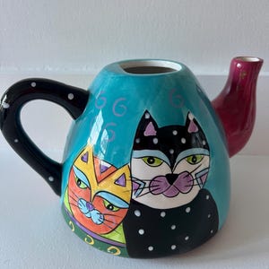 May include: A ceramic teapot with a teal body and a black handle adorned with white polka dots. The teapot features a whimsical design of two stylized cat faces, one orange and yellow, the other black and white. The spout is a deep red color.