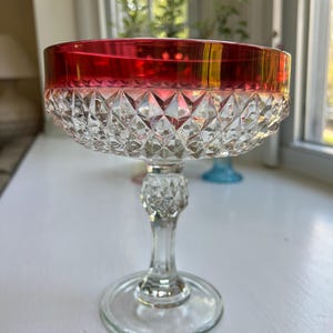 May include: A clear glass champagne coupe with a ruby red rim. The bowl of the glass features a diamond-cut pattern. The stem is clear with a decorative design, and the base is round.