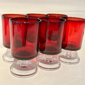 May include: Six red-tinted glass goblets with clear stems and bases. The glasses have a cylindrical shape and are arranged closely together. The goblets are made of a transparent material, allowing light to pass through.