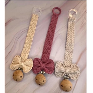 May include: Three crochet pacifier clips in cream and mauve, each with a wooden clip and a bow. The text "Wondie pacifier" is at the top of the image. The clips have a clear plastic loop at the top.