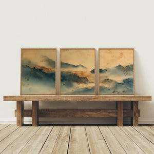 May include: Three framed landscape art prints. Each print features a watercolor-style mountain scene in shades of blue, brown, and yellow. The prints are displayed on a rustic wooden bench against a white wall and wooden floor.