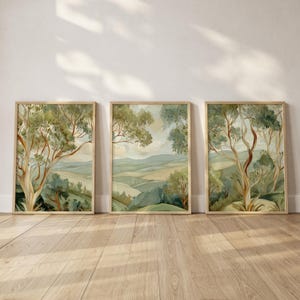 Puede incluir: Tres impresiones artísticas enmarcadas de paisajes. Cada impresión presenta una pintura a la acuarela de una colina verde ondulada con árboles. La obra de arte está en tonos de verde, azul y beige, con un marco de madera clara.