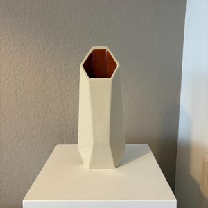 Op de afbeelding: Een geometrische vaas met een gefacetteerde, gebroken witte buitenkant en een gladde, contrasterende terracotta-kleurige binnenkant. De vaas heeft een zeshoekige opening aan de bovenkant en een taps toelopend, hoekig lichaam richting de basis.