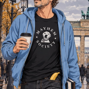 Può includere: Una t-shirt nera con una grafica circolare bianca recante la scritta MAYHEM SOCIETY e l'illustrazione di un cane, indossata sotto una felpa azzurra con zip e cappuccio. L'abbigliamento include jeans scuri con una banana gialla nella tasca.
