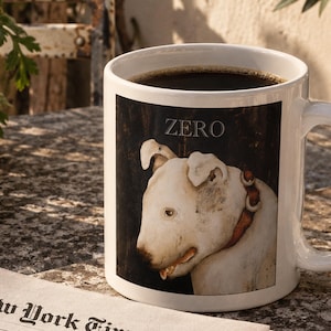 Può includere: Una tazza in ceramica bianca riempita di caffè nero, caratterizzata da una stampa grafica di un bull terrier bianco con un collare marrone. Sopra il cane, la parola ZERO è stampata in lettere maiuscole bianche su uno sfondo scuro.