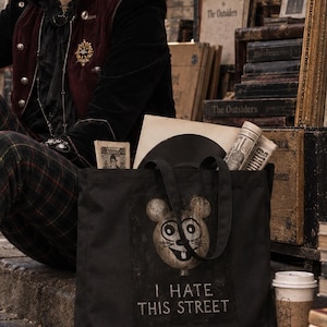 Op de afbeelding: Een zwarte canvas tas met een grafische print van een gestileerde muizenkop met grote ronde ogen en de tekst "I HATE THIS STREET" in witte hoofdletters onder de muisillustratie. De tas is voorzien van twee zwarte stoffen hengsels.