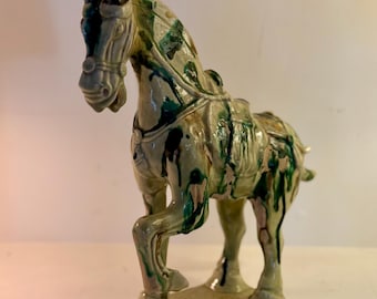 May include: A ceramic horse figurine with a green and cream glaze. The horse is posed with one leg raised, and it has a saddle. The figurine is mounted on a rectangular base.