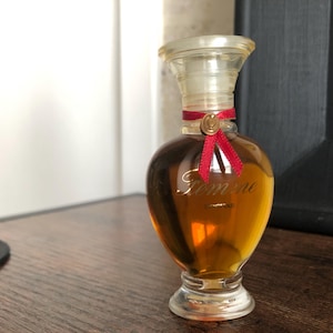 May include: A vintage perfume bottle with a clear glass stopper and a rounded, amber-colored body. The bottle is adorned with a red ribbon and the word "Femme" in gold script. The perfume bottle sits on a wooden surface.