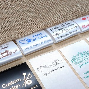 May include: Several white satin labels with printed text and graphics, including "Paws of Love" and "Handmade Design." The labels feature sewing-related imagery and floral designs. The background is a textured, beige fabric, providing a neutral backdrop for the labels.