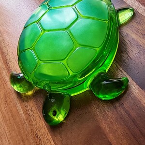 May include: A translucent emerald green glass turtle figurine featuring a detailed shell with raised hexagonal patterns. The turtle has a smooth, polished finish with a distinct head, four flippers, and a rounded back, showcasing a vibrant green hue.
