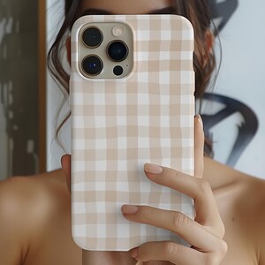 May include: A beige and white gingham phone case. The case is held up in front of a person's face. The case has a square pattern design. The phone case has a camera and flash opening.