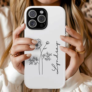 May include: White phone case with a black floral illustration and the name "Lyndsay" in cursive. The case has a glossy finish and a camera cutout. The phone case is held by a person.