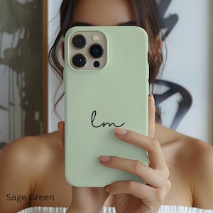 May include: Sage green phone case with a black cursive "lm" design. The case is a solid, light green color and has a smooth finish. The phone case is held up in front of a person.