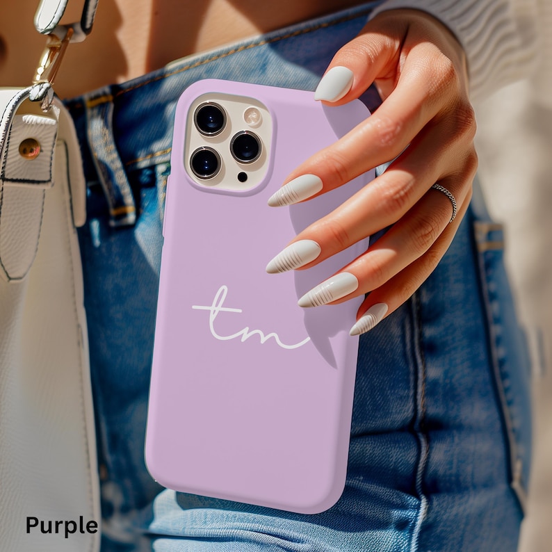 K&ouml;nnte beinhalten: Eine hellviolette Handyh&uuml;lle mit den wei&szlig;en Initialen "tm". Die H&uuml;lle wird von einer Person in der Hand gehalten, wobei die Kamera des Telefons sichtbar ist. Das Wort "Purple" befindet sich unten links im Bild.