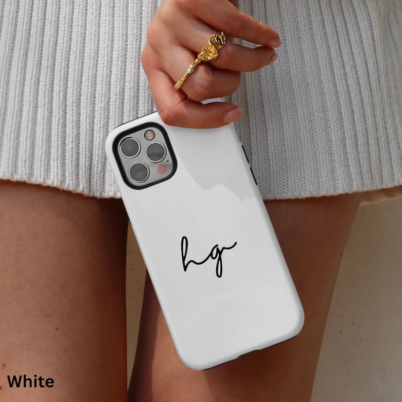 K&ouml;nnte beinhalten: Wei&szlig;es Handycase mit schwarzem Rand und den handgeschriebenen Initialen "hg". Das Case wird von einer Person mit einem goldenen Ring gehalten. Das Handycase ist wei&szlig;.