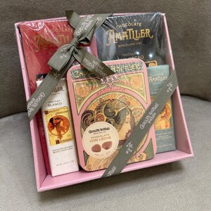 May include: A pink gift box containing Amatller chocolate products. The box includes chocolate bars, a tin of chocolate, and a white chocolate bar. The box is tied with a gray ribbon with the words "Chocolate Amatller Barcelona" printed on it.