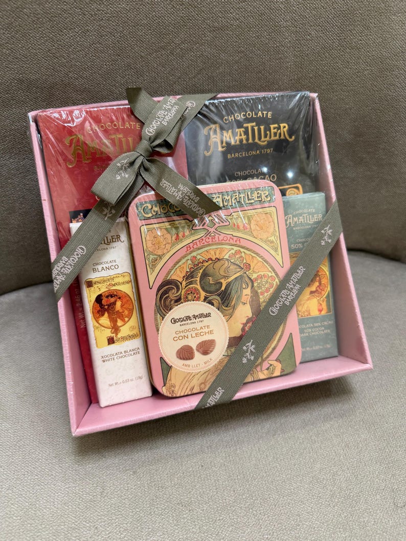 May include: A pink gift box containing Amatller chocolate products. The box includes chocolate bars, a tin of chocolate, and a white chocolate bar. The box is tied with a gray ribbon with the words "Chocolate Amatller Barcelona" printed on it.