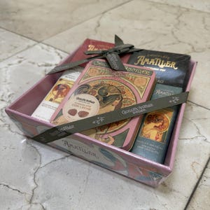 May include: A gift box containing several Amatller chocolate bars and a tin. The box is pink with a floral design and tied with a gray ribbon. The chocolate bars are in various colors and flavors.
