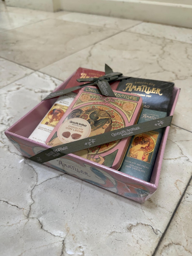 May include: A gift box containing several Amatller chocolate bars and a tin. The box is pink with a floral design and tied with a gray ribbon. The chocolate bars are in various colors and flavors.