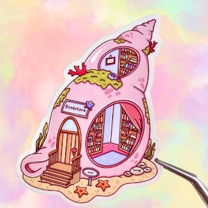 May include: A pink seashell-shaped sticker with a bookstore theme. The sticker features a door, windows, and shelves filled with books. The word "Bookstore" is displayed on a sign. The background is a pastel watercolor wash.