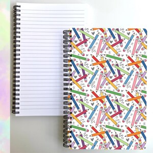 May include: A spiral-bound notebook featuring a white cover with a repeating pattern of colorful pencils in shades of blue, green, yellow, orange, red, and purple, interspersed with small gray hearts and star shapes. The interior pages are white with black lines.