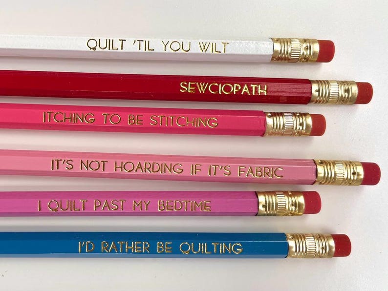 May include: A set of six wooden pencils in white, red, hot pink, light pink, magenta, and blue. Each pencil features a gold-colored metal ferrule, a pink eraser, and gold foil text: QUILT 'TIL YOU WILT, SEWCIOPATH, ITCHING TO BE STITCHING, IT'S NOT HOARDING IF IT'S FABRIC, I QUILT PAST MY BEDTIME, and I'D RATHER BE QUILTING.