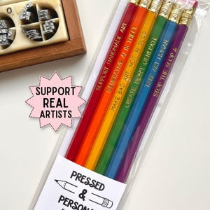 May include: A set of six rainbow-colored pencils in a clear plastic sleeve, each featuring gold-stamped text including SUPPORT HANDMADE ART and GENERATIVE AI IS THEFT. A pink star-shaped sticker reads SUPPORT REAL ARTISTS, and a label reads PRESSED & PERSONAL.