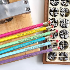 May include: Six hexagonal pencils in pink, yellow, light green, turquoise, light blue, and purple, each with a gold ferrule and pink eraser. The pencils feature the text: DON'T WORRY IT'LL QUILT OUT, MEASURE TWICE, CUT ONCE!, MAKE QUILTING TAX-DEDUCTIBLE, PUT YOUR FOOT DOWN & QUILT, BOBBIN CHICKEN WINNER, and QUILT WHILE YOU'RE AHEAD.